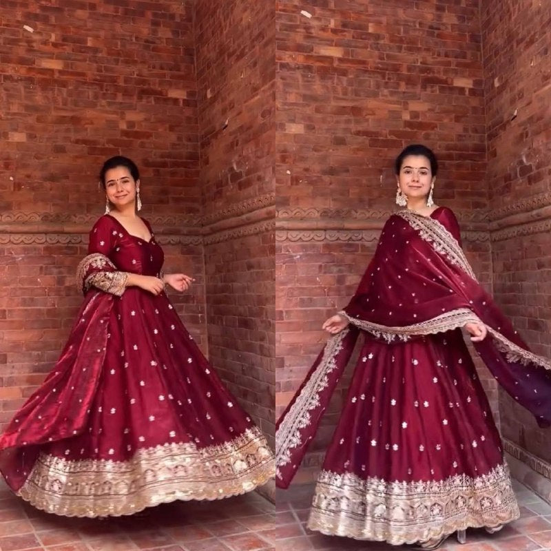 Woman in a maroon traditional outfit with gold details against a brick wall.