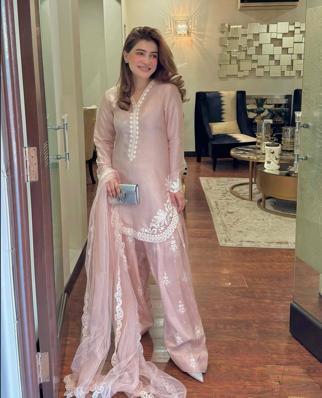 Woman in a light pink traditional outfit with white embroidery in an indoor setting.