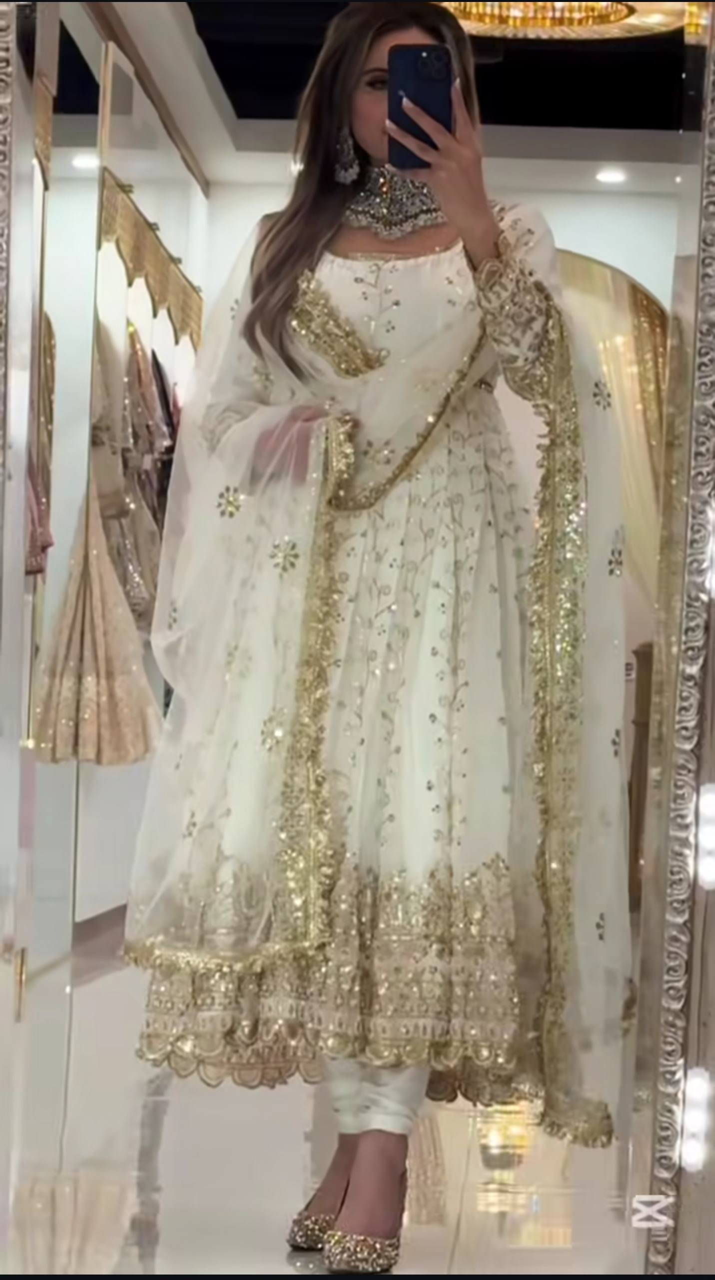 Person wearing a white and gold traditional outfit with a sheer dupatta, standing in front of a mirror.