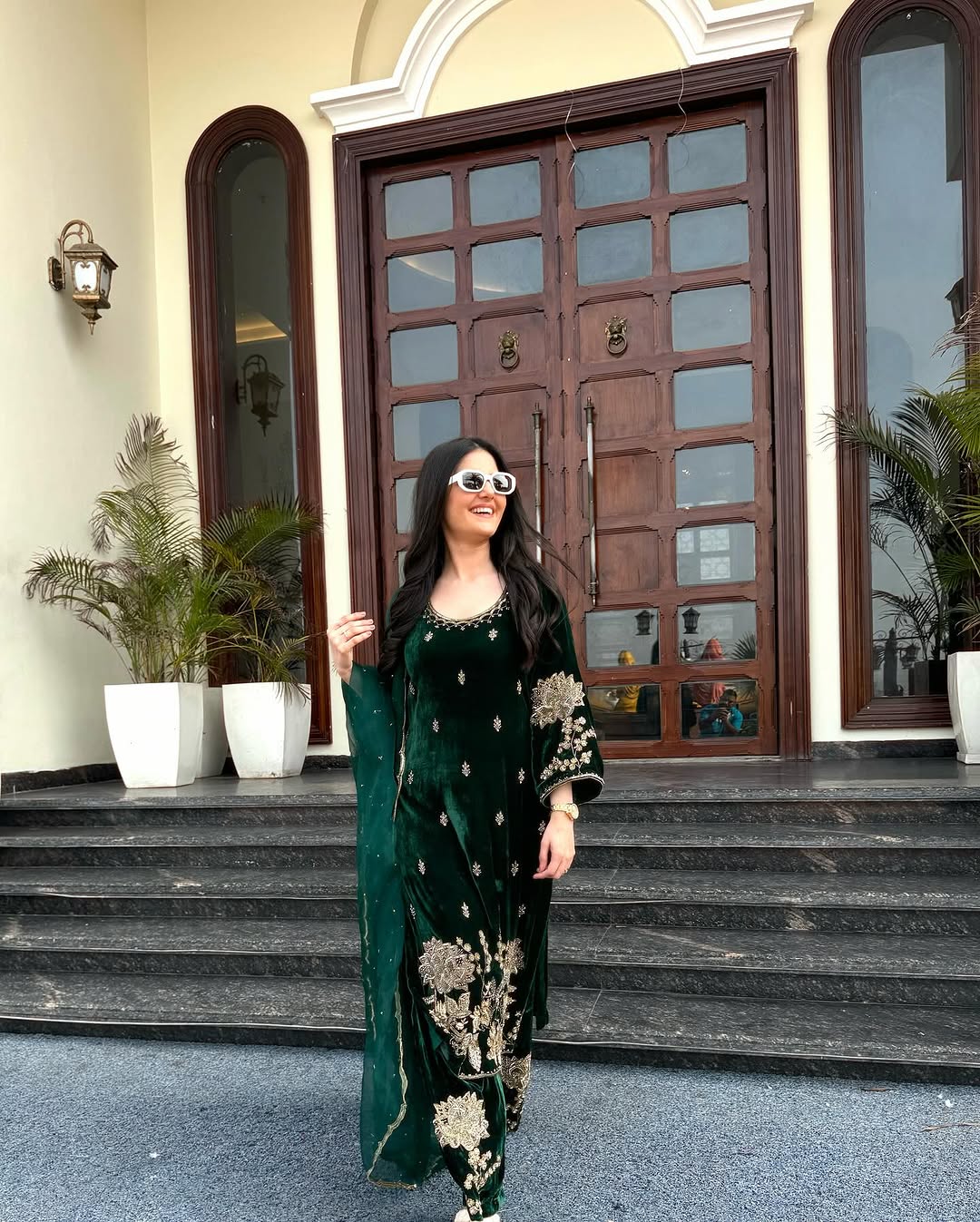 A person standing in front of a large wooden door, wearing a green velvet traditional outfit with gold embroidery and a dupatta.